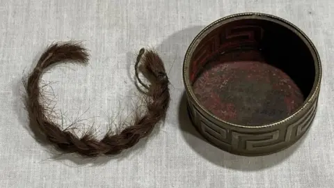 Morgan Evans & Co A dark brown plaited lock of hair is on a piece of cloth on the left hand side of a small gold box. 