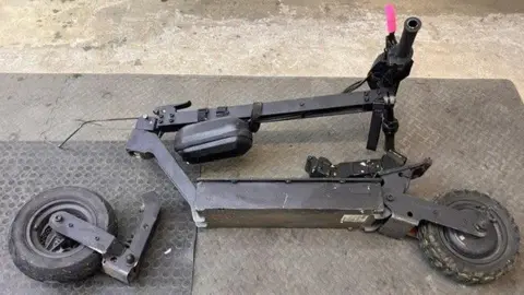 Broken e-scooter pictured on the floor of a repair shop in Aberdeen. The scooter is black with one pink handle. The back wheel and connecting fitting is broken and completely detached from the body of the scooter