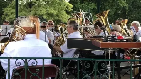 Martin Heath/BBC People sitting in a bandstand holding various instruments including trombones, euphoniums, tubas, cornets and trumpets. The musicians are wearing white shirts and dark trousers. A music stand with clothes pegs attached is visible near the camera, along with a xylophone and a glockenspiel.