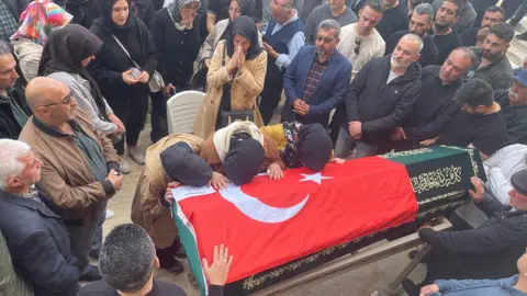 A solemn-looking crowd gathers around a coffin, which has the Turkish flag draped over it, while three women wearing head coverings rest their heads on the coffin.