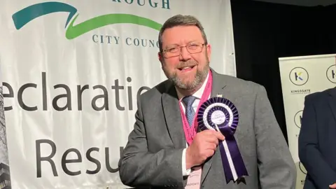 BBC Chris Harper with a beard and glasses wearing a grey suit. He is touching his purple and white rosette with his name on it. A Peterborough City Council sign saying "Declaration Results" is in the background. 