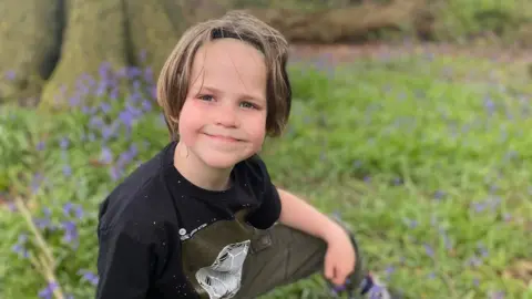 Bev Price A boy with long hair, a black t-shirt and green combat trousers is kneeling in a grassy woodland.
