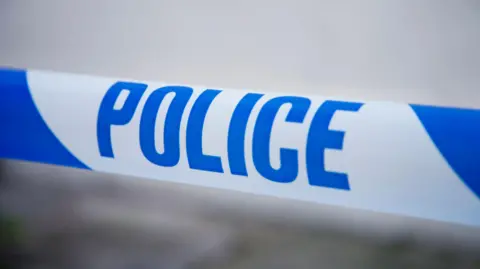 A stock image of some police tape with blue writing on a white background. The background is blurred with no identifiable features.