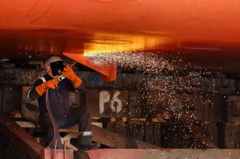 Getty Images A man in a blue boiler suit cuts off a metal stay from beneath a large ship