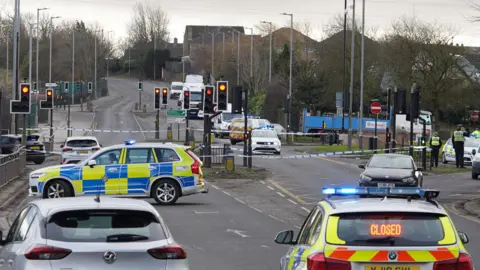 A two lane room with a number of police vehicles blocking the carriageway and police tape stretched across the road