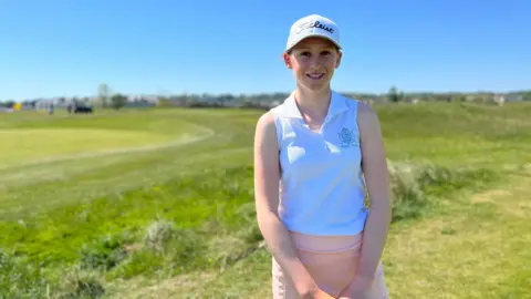 Anabelle Lucas-Villar smiles at the camera as she wears a pink skirt and white sleeveless top and a white cap. Behind her is a golf course.