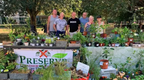 A group of six people (four men, two women) stand behind tables of plants at a plant sale to raise money for the Preserve Littleworth Lane Fields campaign. A banner on one of the tables says 'Preserve Littleworth Lane Fields