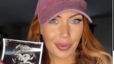Hayley Pearce Hayley is holding her baby scan picture and smiling at the camera, She has long red wavy hair and is wearing a pink cap. 