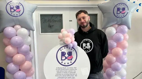 BBC Paris Theodorou is surrounded by balloons at the launch of Blanket Bank. He has short dark hair and a dark beard and wears glasses. He has a black hoodie on with the words "Blanket Bank" in a circle on it and stands behind a circle with writing about the bank on it. Either side are swirls of lilac, pink and white balloons and he stands in front of white doors.