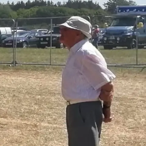 PA Media An old man wearring suit trousers and shirt, and a bucket hat, standing side on in a field.
