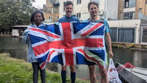 A Future Without Rubbish A woman and two men on a canal side in jogging gear and gloves, with one holding a rubbish bag and all standing behind a large Union Jack which is draped across them.