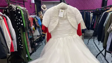 A large white dress is seen hung up on a clothes rail in a purple and black room.
There are a number of clothes on rails on either side of the dress.