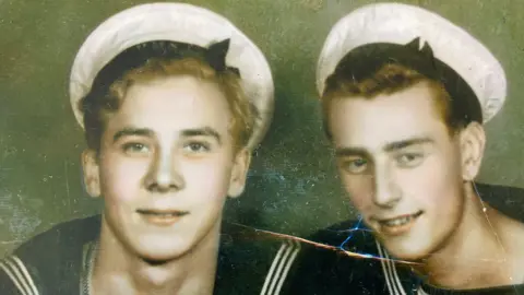 Ken Foster Old, crackled picture of Ken when a teenager in a sailors hat and uniform next to another young man in the same outfit. Both smiling at the camera.