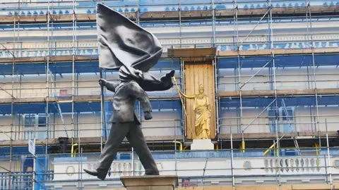 Statue of a suited figure holding a flag, seen against a scaffolding‑covered building in London