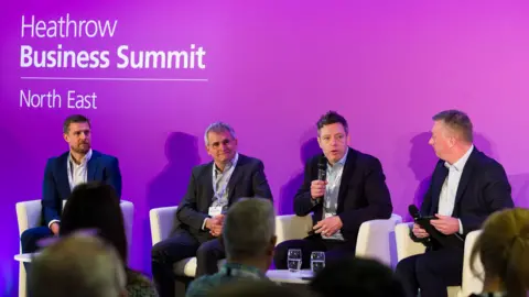 Supplied Four men with short hair wearing light shirts and dark suits, sat on white chairs with two of them holding microphones. There is a purple background and two glasses on a white table. 