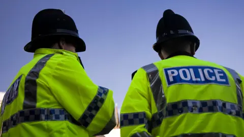 Two police officers wearing uniform stand next to each other with their backs facing the camera.