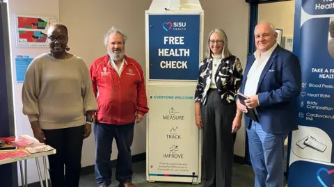 Guy Campbell/BBC Four people stand beside the 7ft high white machine which has sign on it reading "Free Health Check - Everyone Welcome"