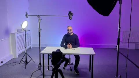 SuperSaf producing content in a bare room with lighting equipment and video camera