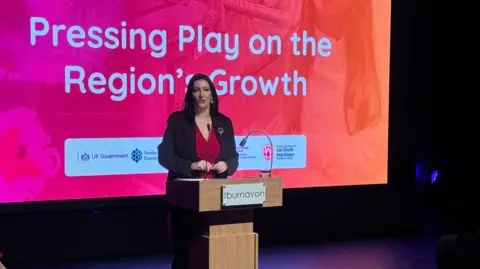 A lady in a black blazer and red top, stands talking into a microphone at a wooden plinth. She has dark brown hair.