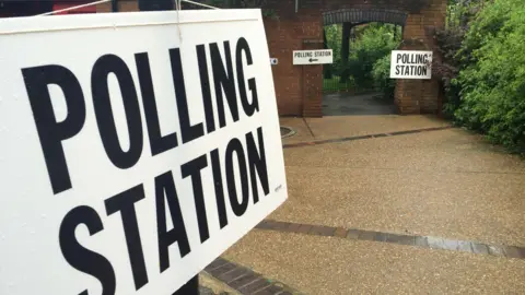 A generic image of a white sign with black lettering for a polling station. In the background there is an arched entrance to a building's garden with further signs pointing to a polling station.