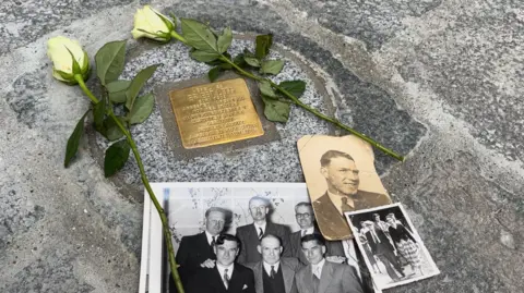 A commemorative brass plaque and grey stone. Aged photos of Legg and others, along with two white roses lay atop the stone.