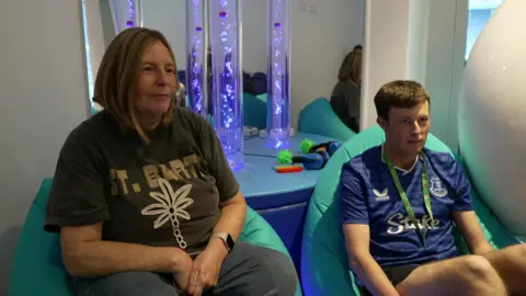 Julie and Joe Simpson are sitting on bean bags in the sensory room at Everton's new stadium. Tubes filled with blue/purple liquid and bubbles can be seen behind them. They are both smiling. Joe is wearing an Everton shirt and shorts and a sunflower lanyard.