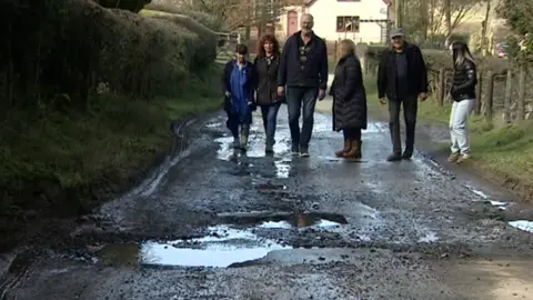 Six Cleeton St Mary residents walk down a potholed-road. A hedge runs along one side and a fence on the other. The residents are in the distance with the potholes in the foreground.