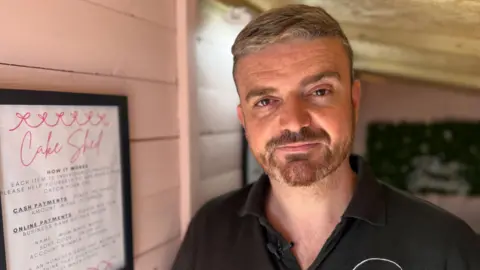 BBC News Lee McCann - a man with short light coloured hair combed over to the left - looks at the camera.
He has a worn expression on his face. He is standing in a small shed, with wooden walls painted pink.