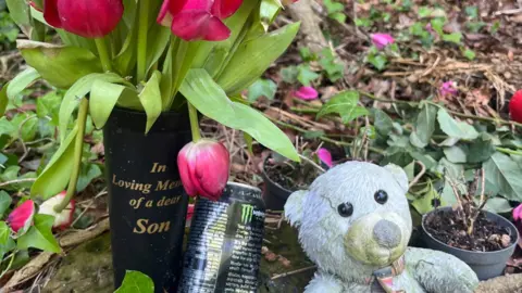 Family handout A black vase containing red flowers with a can of energy drink and a grey teddy bear is lain on a floor covered in ivy and leaves. The case says "In Loving Memory of a dear Son".