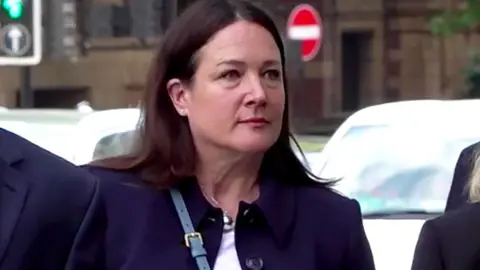 A woman arrives at court wearing a black coat and white top, she has mid length dark brown hair