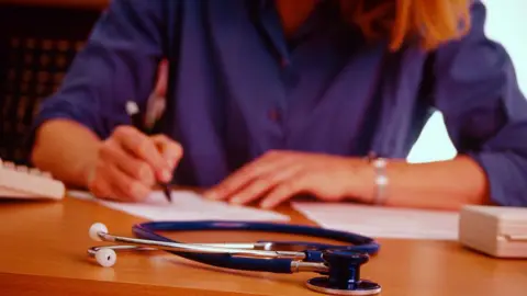 Getty Images A woman with blonde hair and a blue shirt is sitting at a desk and writing. In front of her on he desk is a stethoscope
