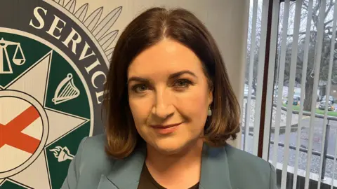 Detective Superintendent Kerry Brennan has shoulder length brown hair and is wearing a blue blazer with a black top. Behind her is the PSNI badge printed on the wall.