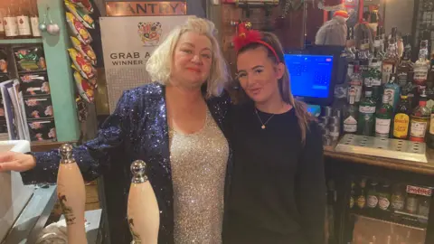 Justin Dealey/BBC Niki Hackett is dressed in a blue glittery cardigan and a silver glittery top. She is stood behind the bar with a younger member of staff dressed in black and wearing a Christmas themed headband.