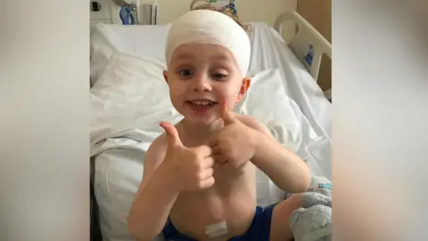 Robinson family A young boy sits in a hospital bed with a bandage on his head. He smiles and puts both thumbs up to the camera.