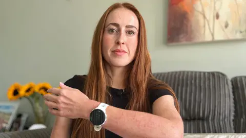 Emma Baugh/BBC Hannah Stephenson is sitting on a grey sofa in her living room. She has long auburn hair which is down, and she is wearing a black short-sleeved T-shirt and sports watch on her wrist. 