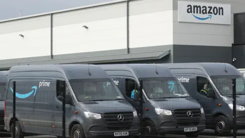 Amazon depot with three vans parked in the foreground with their Amazon livery on a grey background.
