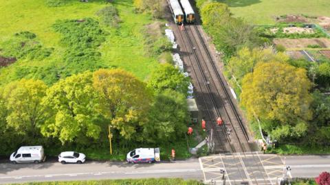 An aerial image of two stopped trains side by side ahead of a level crossing in a rural area surrounded by trees