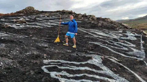 Allie Evans Dean Saunders in a blue top and blue shorts on a blackened mountain in yellow trainers