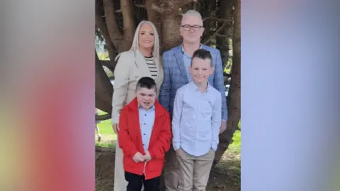 Family handout Alfie (bottom left) with his family. A woman behind him has blonde hair, wearing a beige coat and black and white striped top. A boy beside him is wearing a a light blue shirt, beige chinos and blue trainers. Alfie is wearing a red coat, light blue shirt, black trousers and brown shoes. His dad Jamie is wearing black glasses, a blue check blazer and blue shirt. They are all standing in front of a tree.