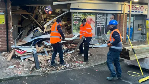Qays Najm/BBC Three people are working to clear the rubble away from the Tesco Express store. They are all wearing orange fluorescent jackets and vests. 