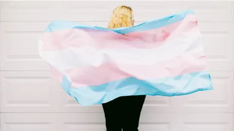 DBenitostock/Getty Images Transgender person from behind, wearing pink and white striped sweatshirt, holds transgender flag