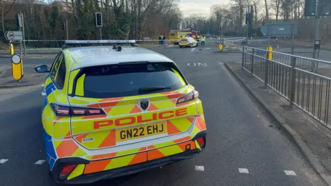 The back of a sign-written police car blocking a road with cordons in place in the distance