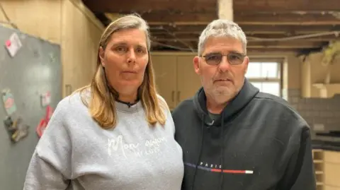 Geraldine Castell wearing a grey jumper with mid-length blonde hair standing next to Carl Castell also wearing a black jumper with their former kitchen in the background