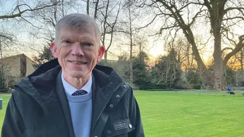 Steve Hubbard/BBC David Day, who has white short hair. He is wearing a blue jacket over a lighter blue jumper, white shirt and blue spotted tie. Behind him is a lawn and across it is a large London plane tree. 