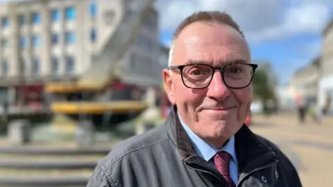 Tudor Evans, Labour leader on Plymouth City Council, in a blue shirt and burgundy spotted tie with a grey jacket and wearing glasses, in Armada Way in Plymouth.