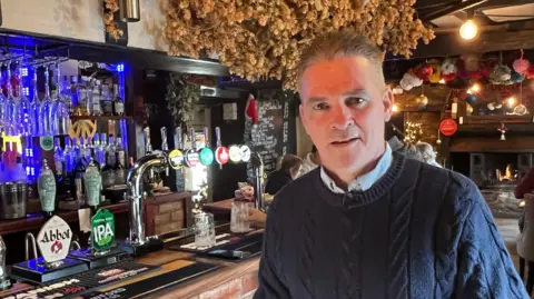 A middle-aged man in a blue jumper leaning against a bar with beer taps in the background. There are beer hops hanging from the ceiling.