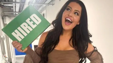 Nadia Suliaman Nadia Suliaman, has a brown bardot dress on and smiles looking up to the ceiling holding a green sign reading "hired" in large white text. She has dark brown hair and brown eyes. 