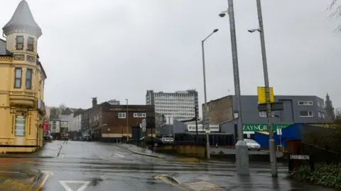 Iconic Media Street view of Heatley Street where the yellow bus gate sign on a lamp post to the right has been turned around. It is on a wet crossroads with a grey sky and a mixture of old buildings in the distance.