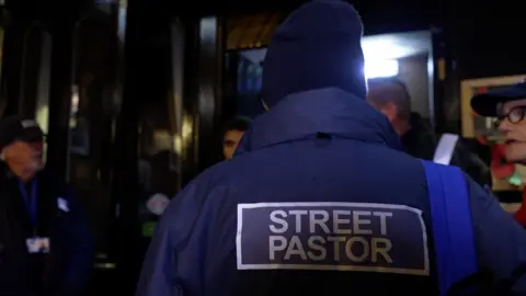 A person in a blue jacket with "street pastor" on the back, talking to a group of people at night in Stafford town centre.