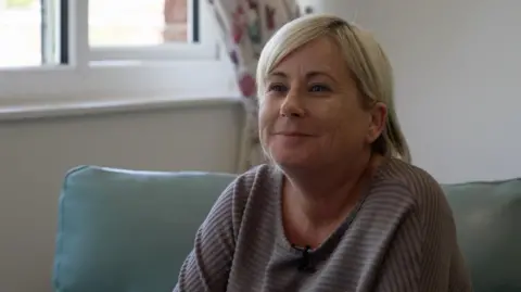 Donna sits on a sofa and smiles. She has a long-sleeved purple top on and her hair is styled in a blonde bob.